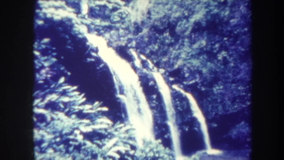 1967 Honolulu home-movie frame of a bright tropical waterfall dropping through dense forest vegetation into a shaded pool.