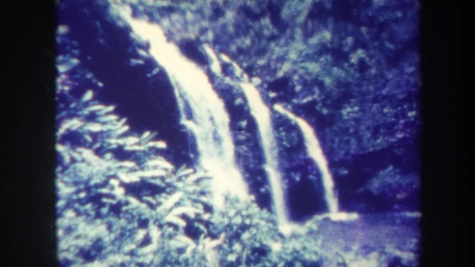 1967 Honolulu 8mm frame showing a tropical waterfall, leafy plants, dark rock, and a small pool in a forested ravine.