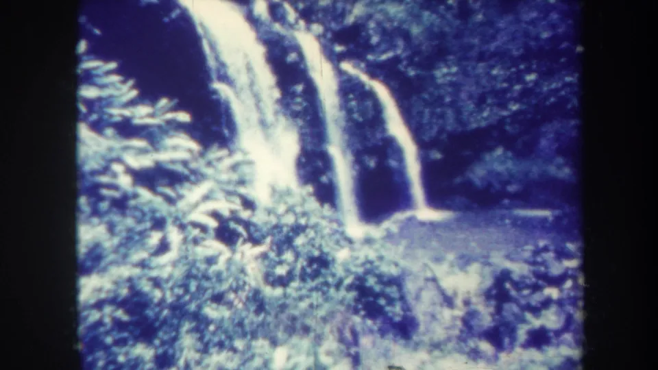 1967 Honolulu home-movie frame with a blue-tinted waterfall, lush green plants, rocky cliff, and pool at the base.