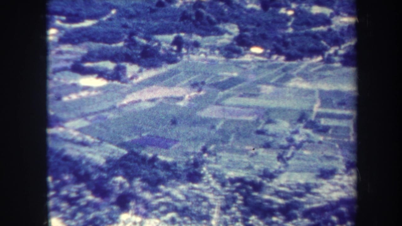 Aerial view of fields, trees, and scattered buildings in a blue-tinted 8mm frame.
