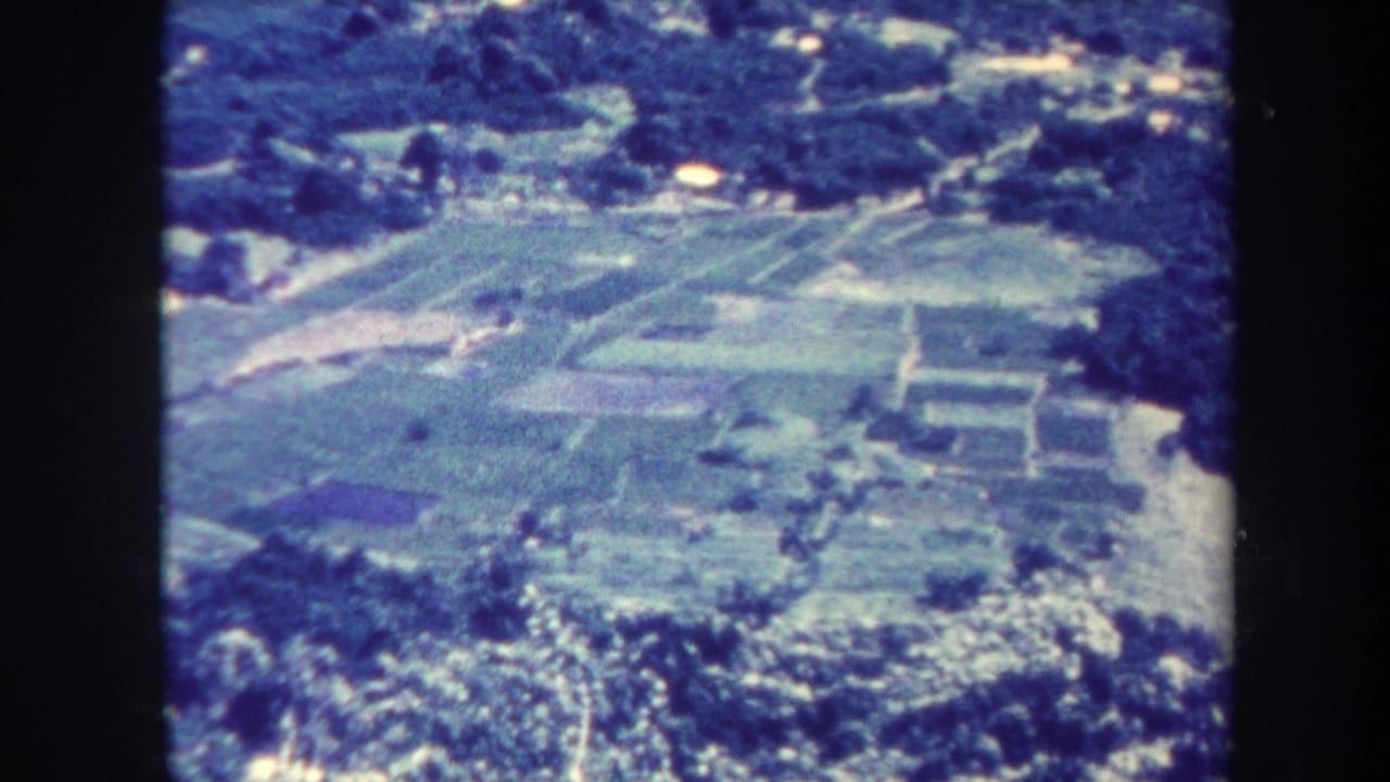 Aerial landscape with farmland, trees, and pale buildings seen through aged film grain.