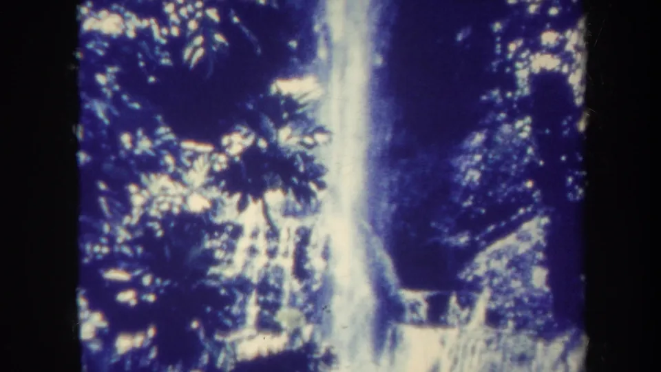 1967 Honolulu 8mm frame showing a steep waterfall partly framed by dense tropical leaves and shadowed rock.