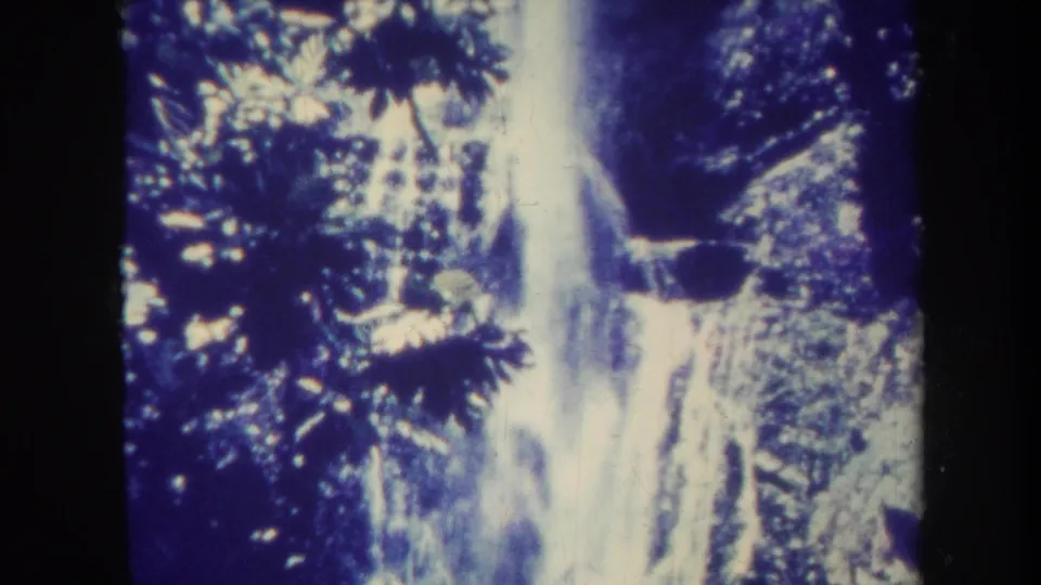 1967 Honolulu 8mm frame with white water falling down a steep, shadowed rock surface surrounded by tropical vegetation.