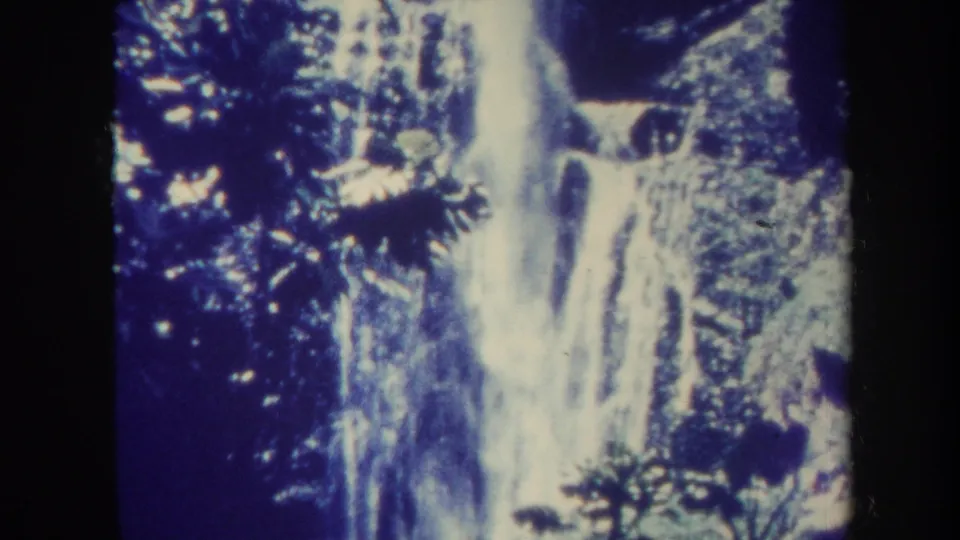1967 Honolulu 8mm frame showing the waterfall continuing over rocks, with leafy plants and dark forest framing the scene.