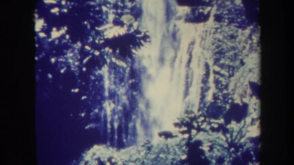1967 Honolulu 8mm frame showing a steep waterfall, rocky slope, and dense tropical growth in blue-shifted archival color.