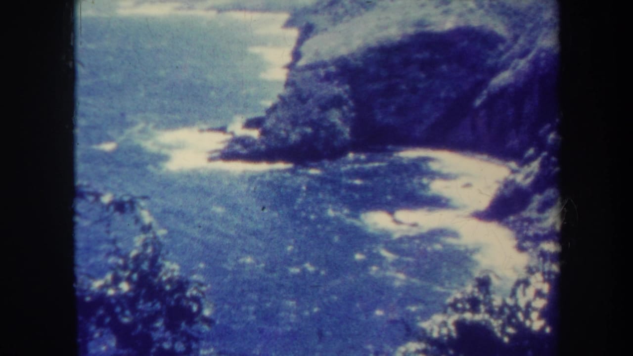 1967 Honolulu 8mm frame of a rugged coastal shoreline with blue ocean, pale sand coves, white surf, and vegetation-covered cliffs.