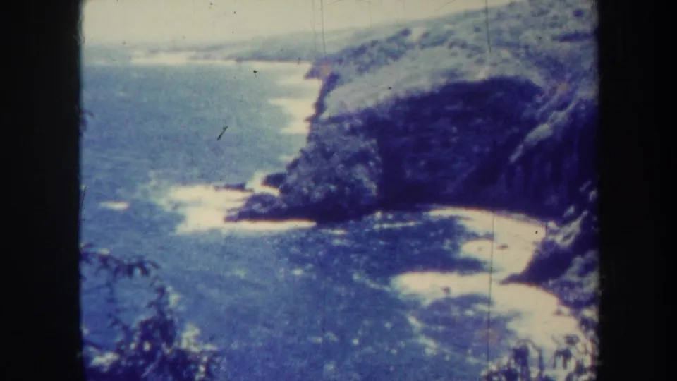 1967 Honolulu 8mm frame showing a slightly washed coastal view with surf lines, pale beach sand, rocky hillside, and foreground vegetation.
