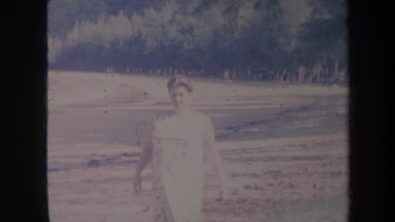 Woman in a light dress standing on an open plain with a wooded hillside behind her.