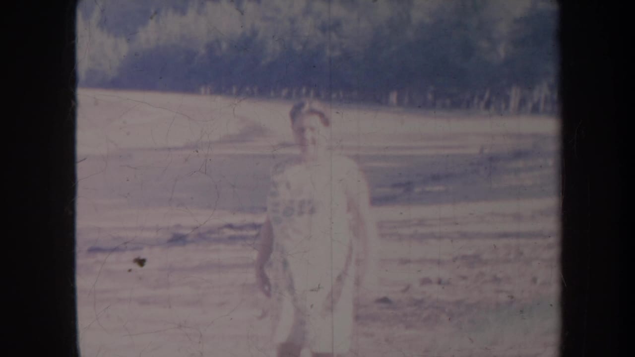 Woman standing outdoors on pale open ground with trees and a fence-line in the background.