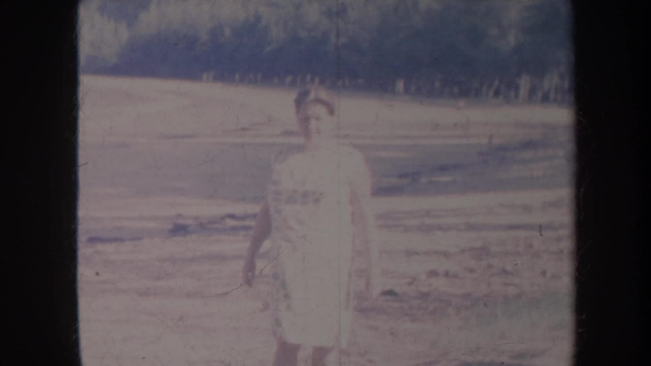 Faded 1967 Honolulu home-movie frame showing the woman slightly lower in view, with open land and a dark wooded hillside behind her.