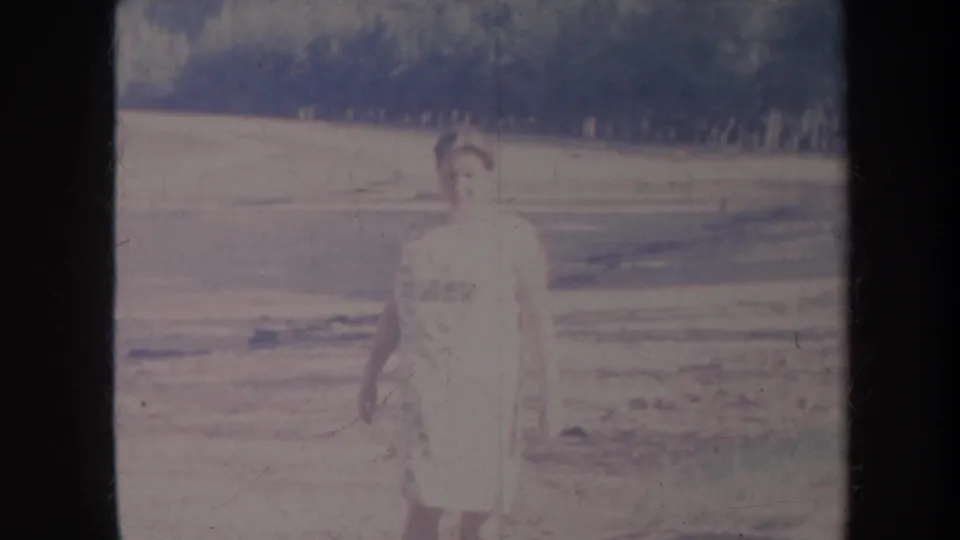 Faded 1967 Honolulu home-movie frame showing the woman slightly lower in view, with open land and a dark wooded hillside behind her.