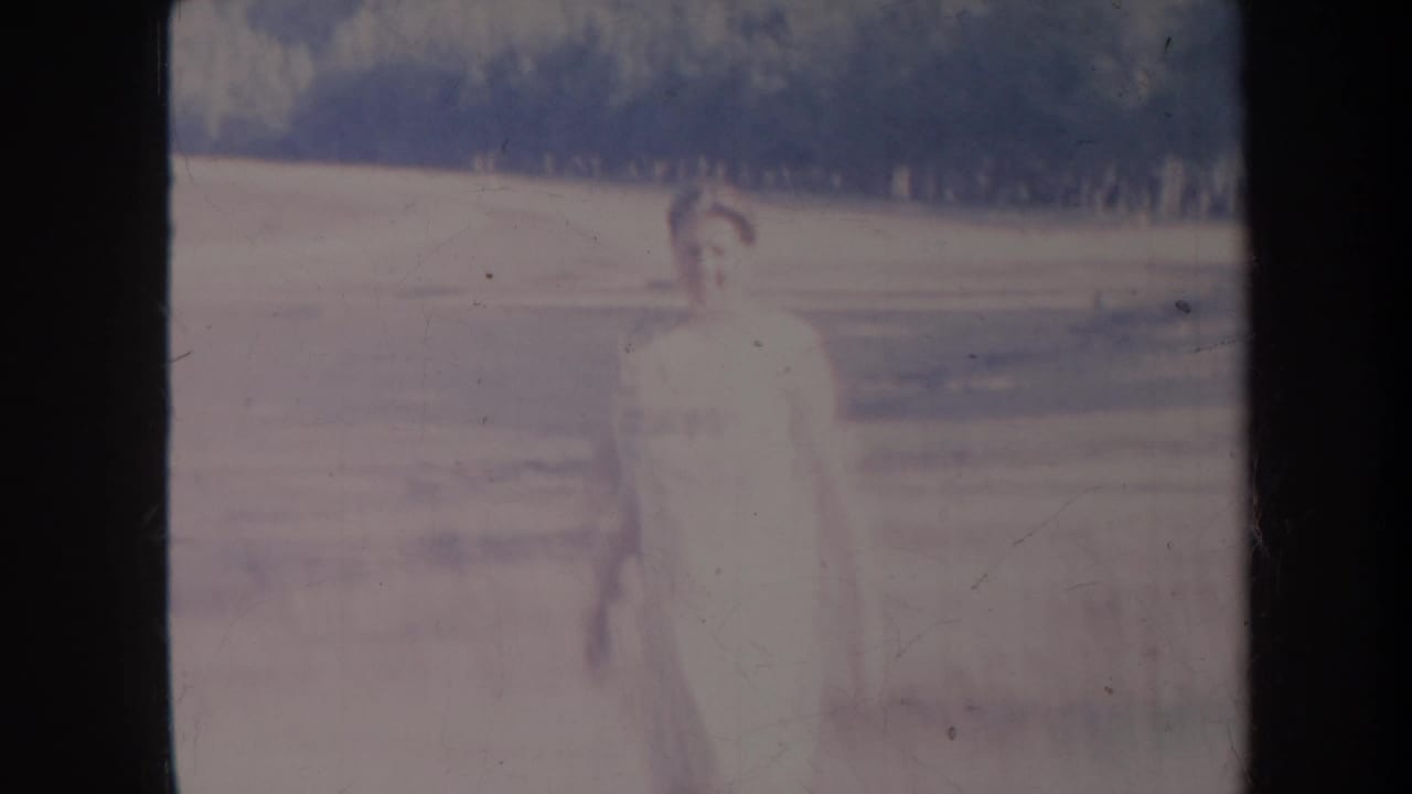 1967 Honolulu 8mm frame with the woman centered in a washed, softly focused outdoor landscape before a distant tree line.