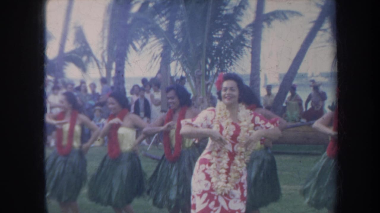 1967 Honolulu home-movie frame showing hula dancers in leis and skirts performing near a beach with palms and spectators in the background.