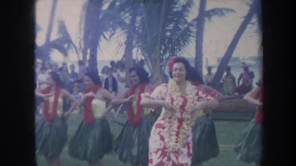 1967 Honolulu home-movie frame showing hula dancers in leis and skirts performing near a beach with palms and spectators in the background.