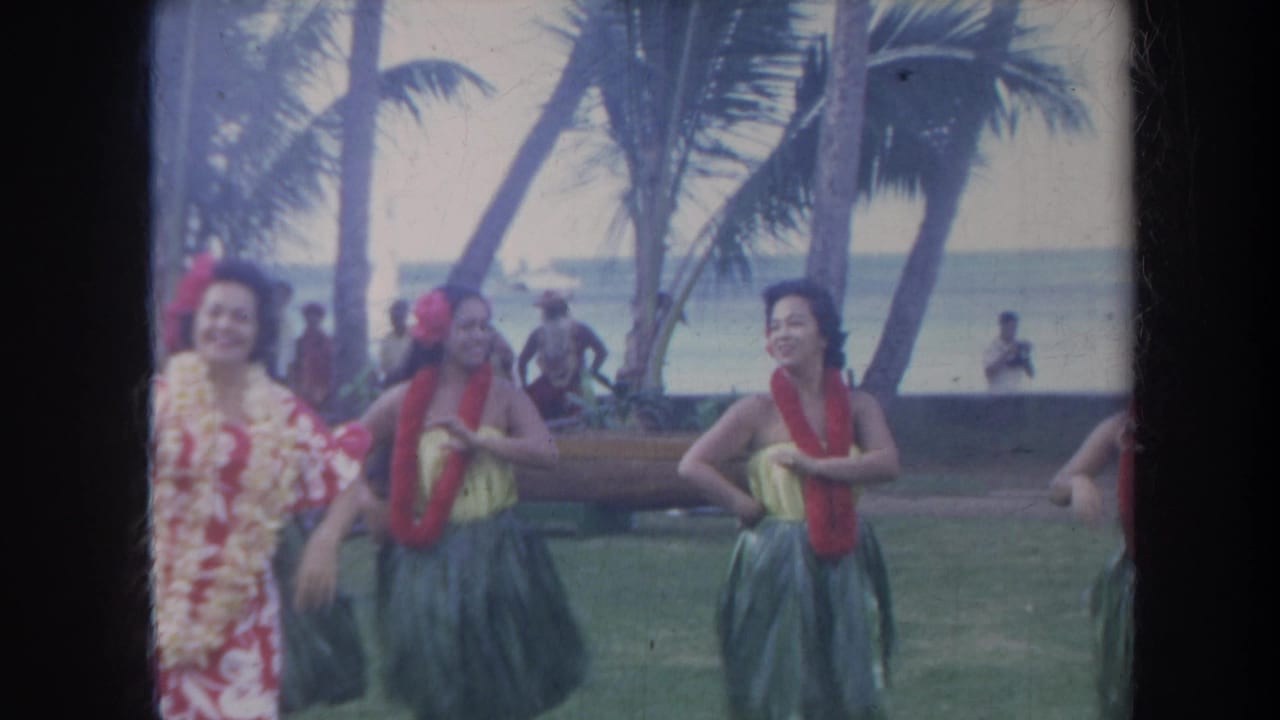 Women in leis and skirts dance near palm trees with the ocean visible beyond them.