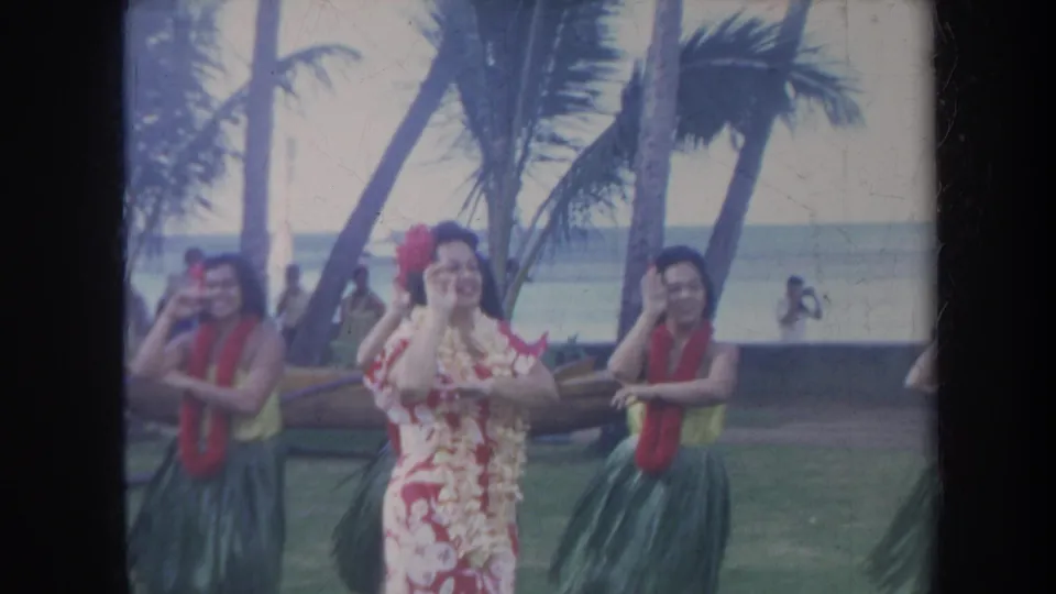 1967 Honolulu home-movie frame with a central dancer in a red and white floral dress framed by hula dancers in leis near the ocean.