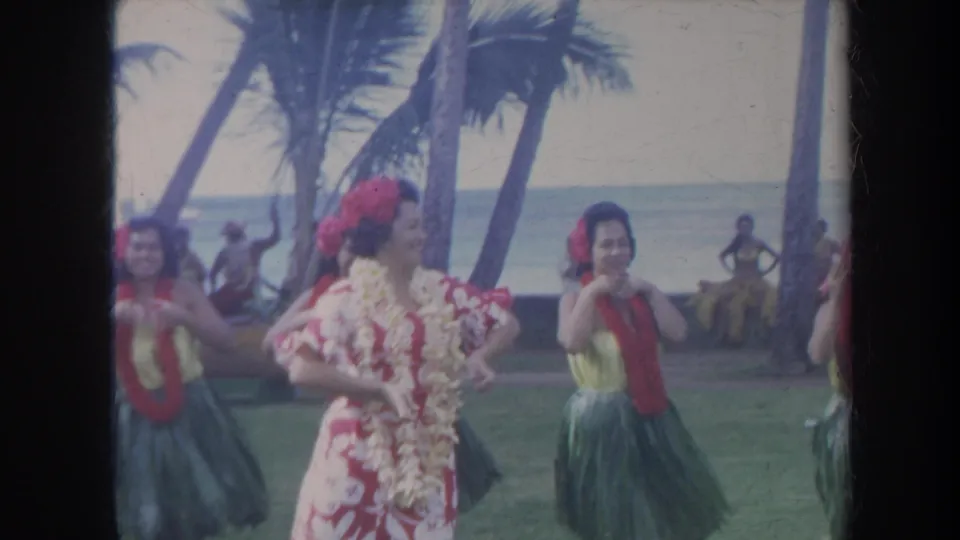 1967 Honolulu frame showing a floral-dressed hula performer turned in profile, with dancers, palm trees, ocean, and onlookers behind her.