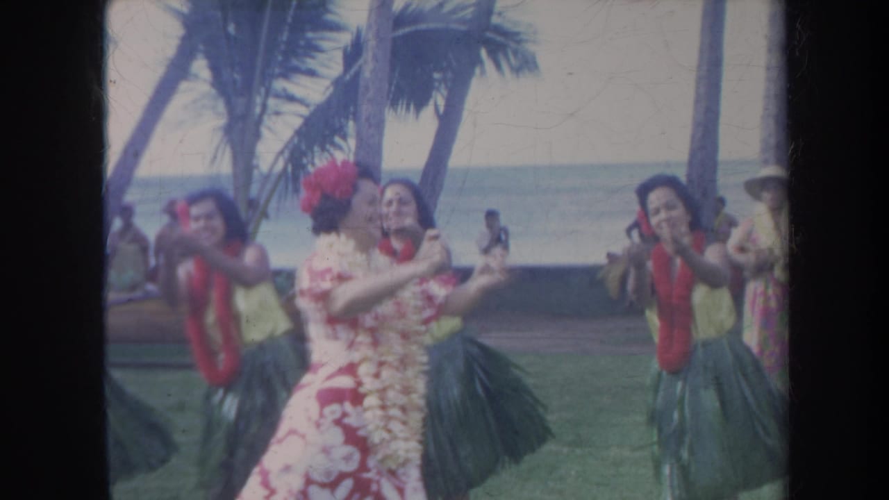 1967 Honolulu home-movie frame showing hula dancers and a floral-dressed performer dancing on grass beside palms and the sea.