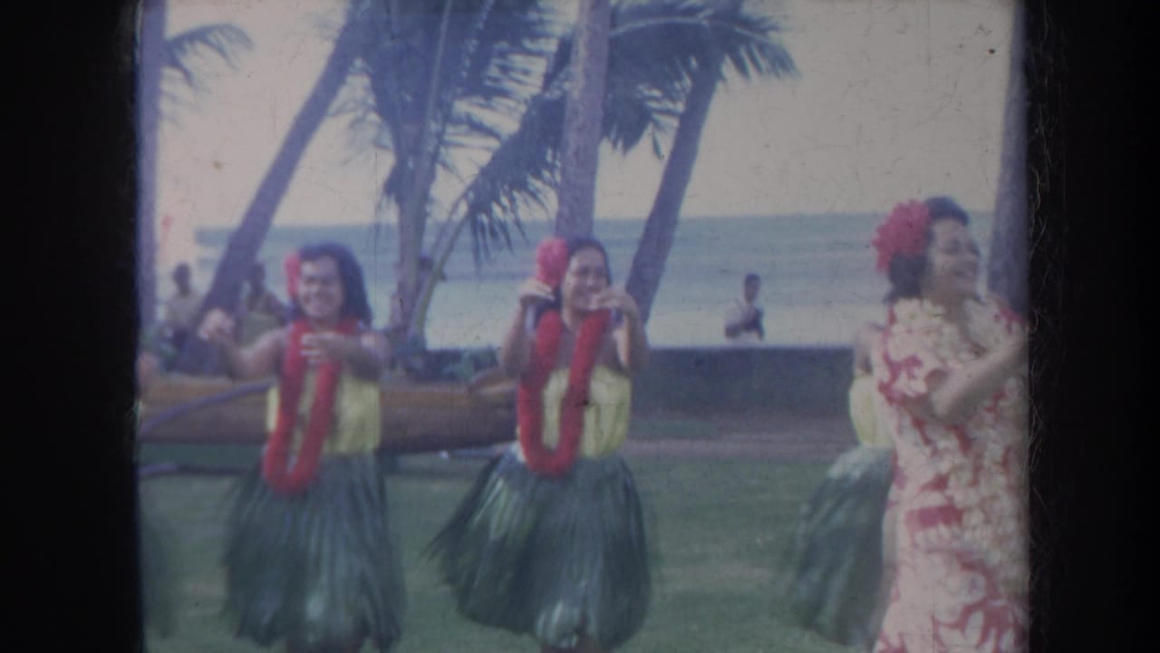 Two hula dancers face the camera while a floral-dressed dancer moves at the edge of the frame.