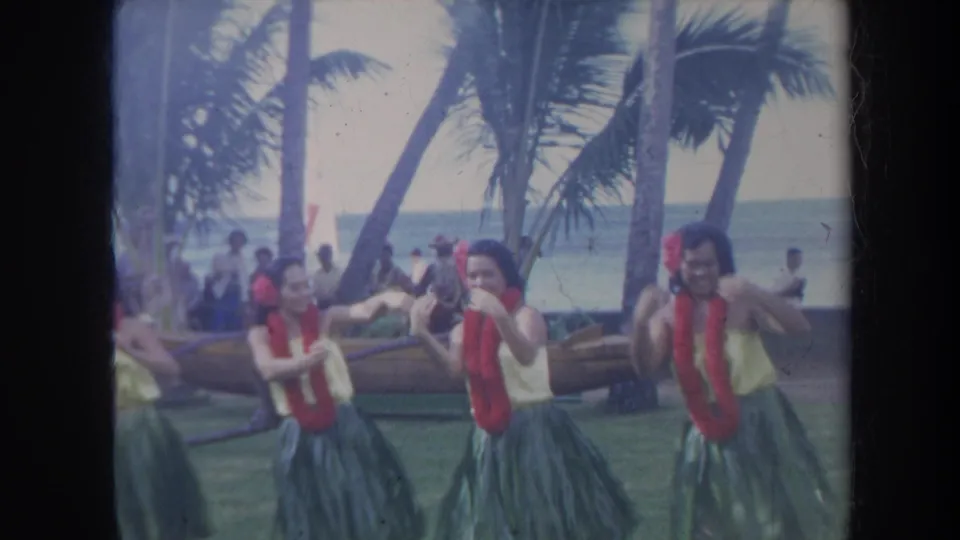 Honolulu 1967 home-movie still of hula dancers in grass skirts and red leis moving together near the beach.