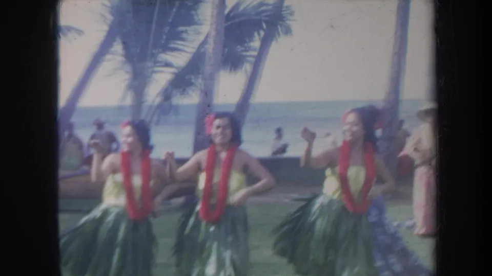 Soft 8mm frame from 1967 Honolulu showing hula dancers in leis and grass skirts performing outdoors by the ocean.