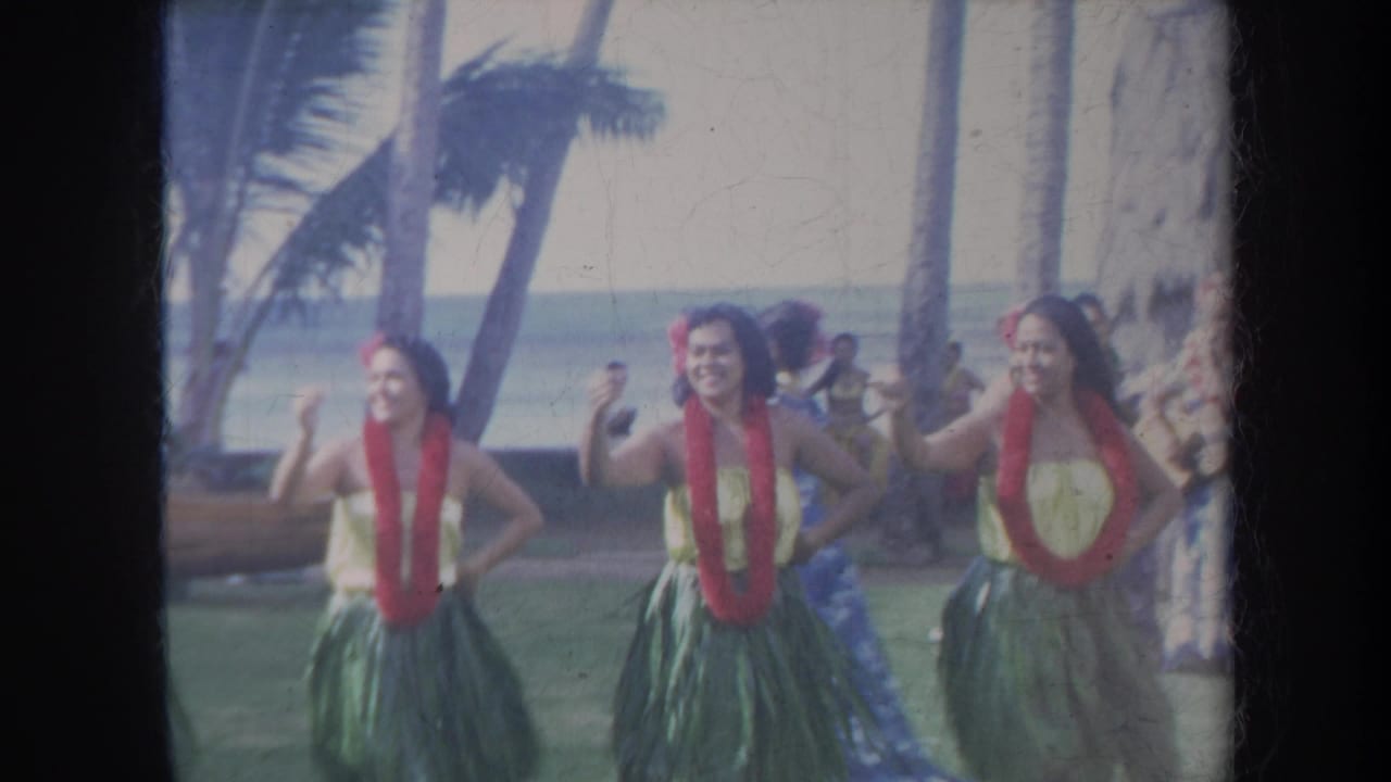 Three hula dancers in leis and grass skirts perform near palm trees by the ocean.