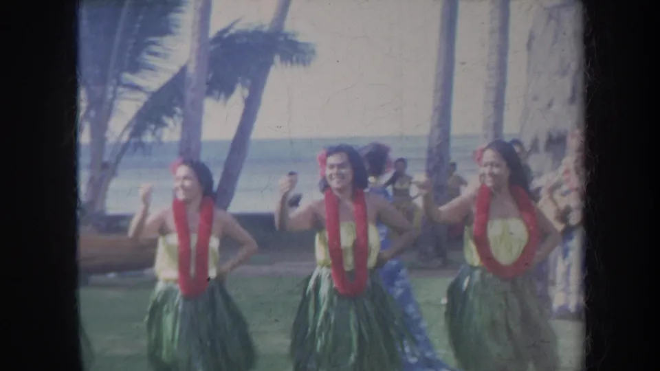 1967 Honolulu home-movie frame showing hula dancers in red leis and green skirts performing near palm trees with the ocean behind them.