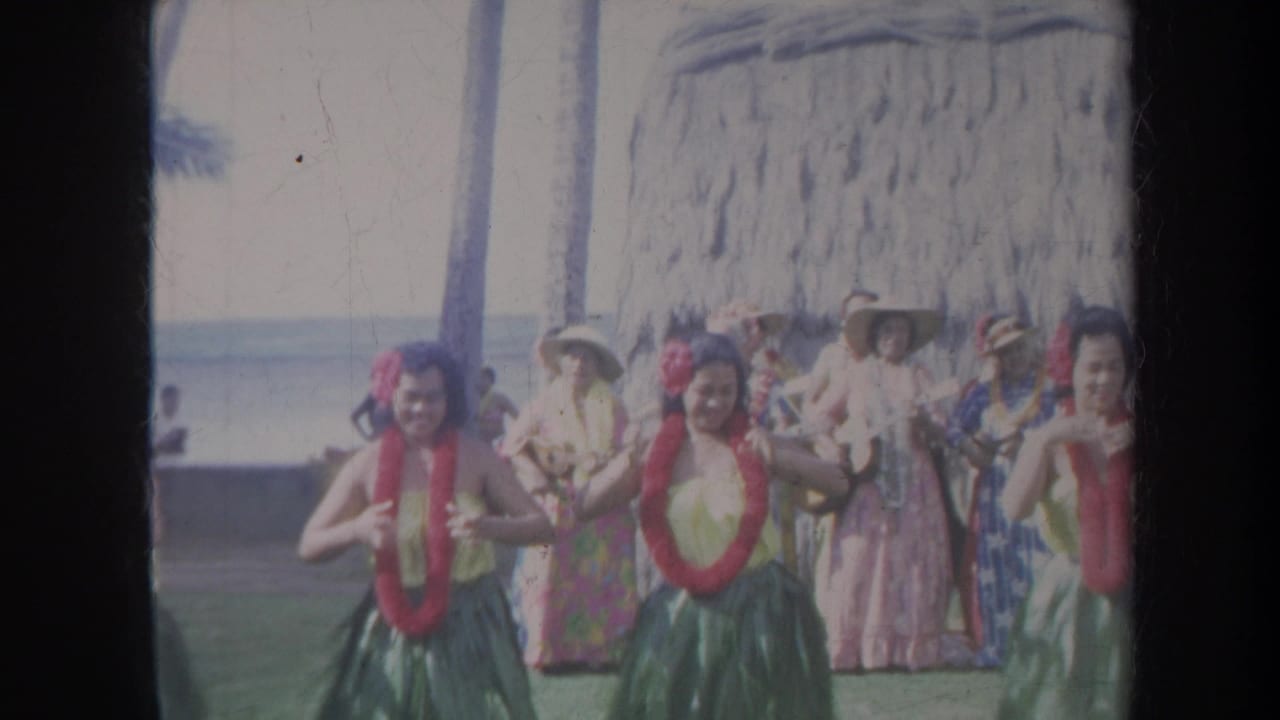 Hula dancers perform in front of spectators near a thatched beach structure.