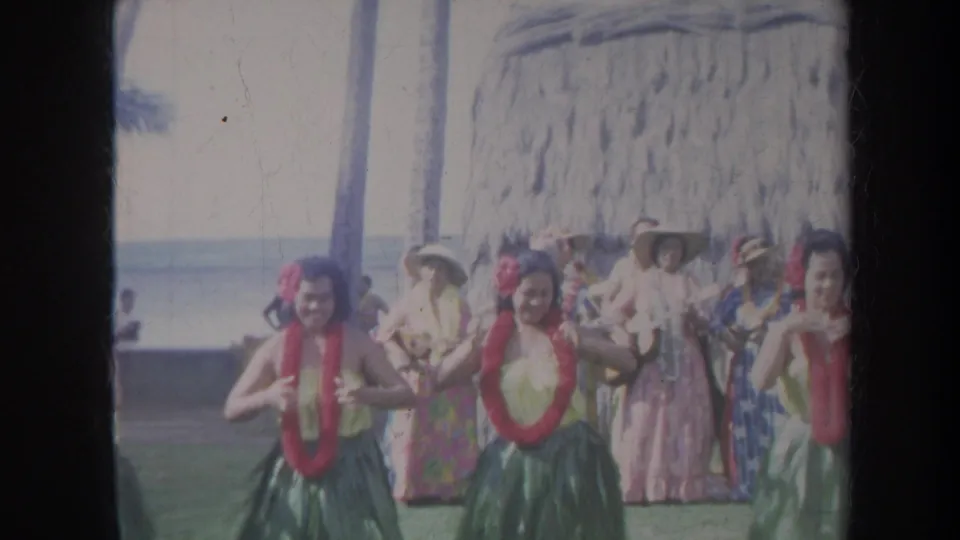 1967 Honolulu frame with hula dancers in red leis performing as spectators in dresses and hats watch near a thatched structure.