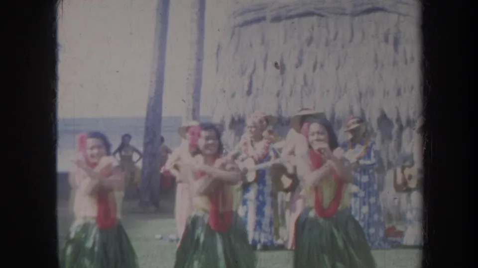 1967 Honolulu home-movie frame showing dancers moving in unison, with spectators and a thatched structure in the beachside background.