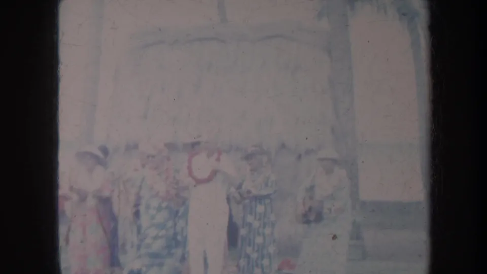 Faded 1967 Honolulu 8mm frame with dancers and spectators still visible near a thatched beach structure and ocean backdrop.