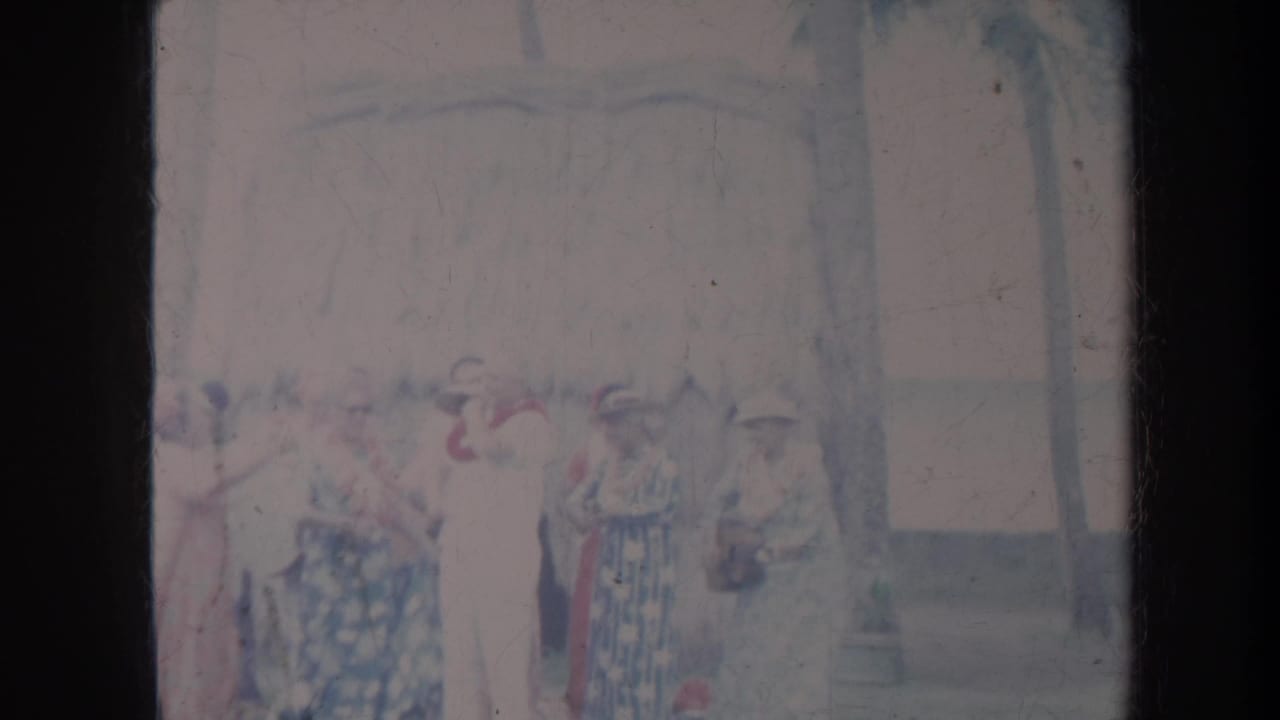 Women in dresses and hats gathered outdoors beneath palm trees and a thatched roofline.