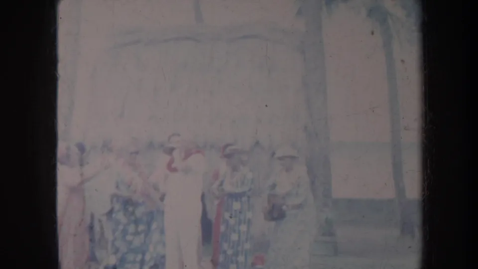 1967 Honolulu home-movie frame showing women in floral dresses and hats standing outdoors near palm trees and a thatched structure.