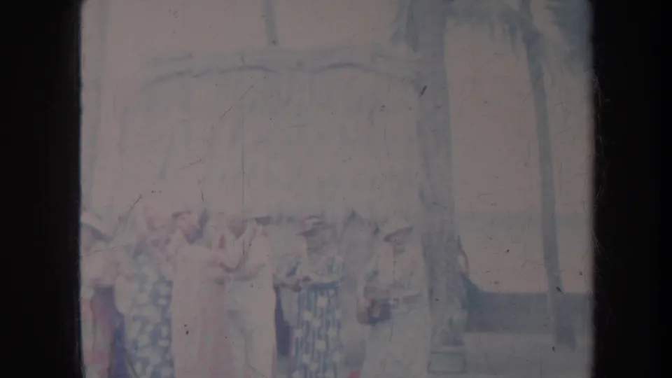 1967 Honolulu frame with women grouped outdoors, including a central woman in white and red, with palms and a shaded roof behind them.
