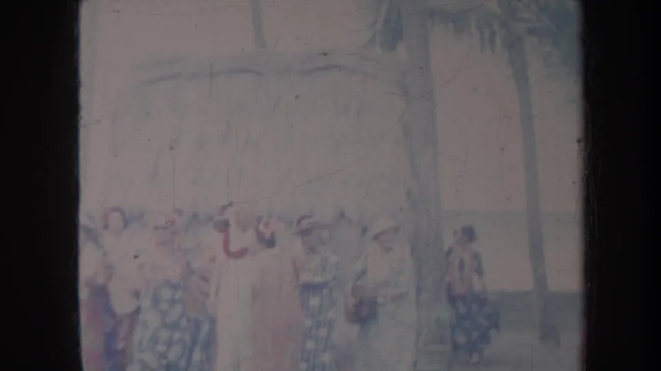 Faded 8mm frame from Honolulu showing women in floral dresses gathered outdoors with palm trees and a shaded roofline visible.