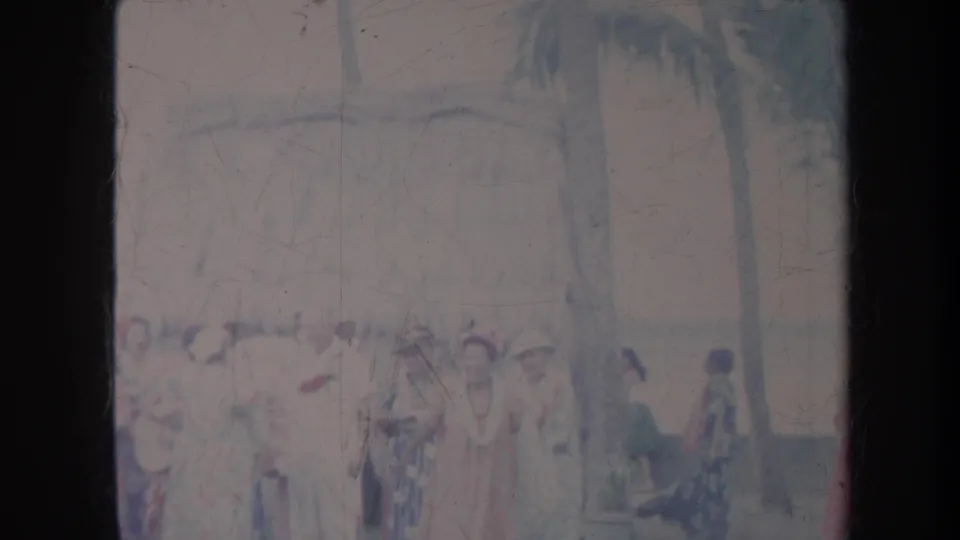 1967 Honolulu home-movie frame showing women in dresses near palms, with a seated person at the right side of the outdoor scene.