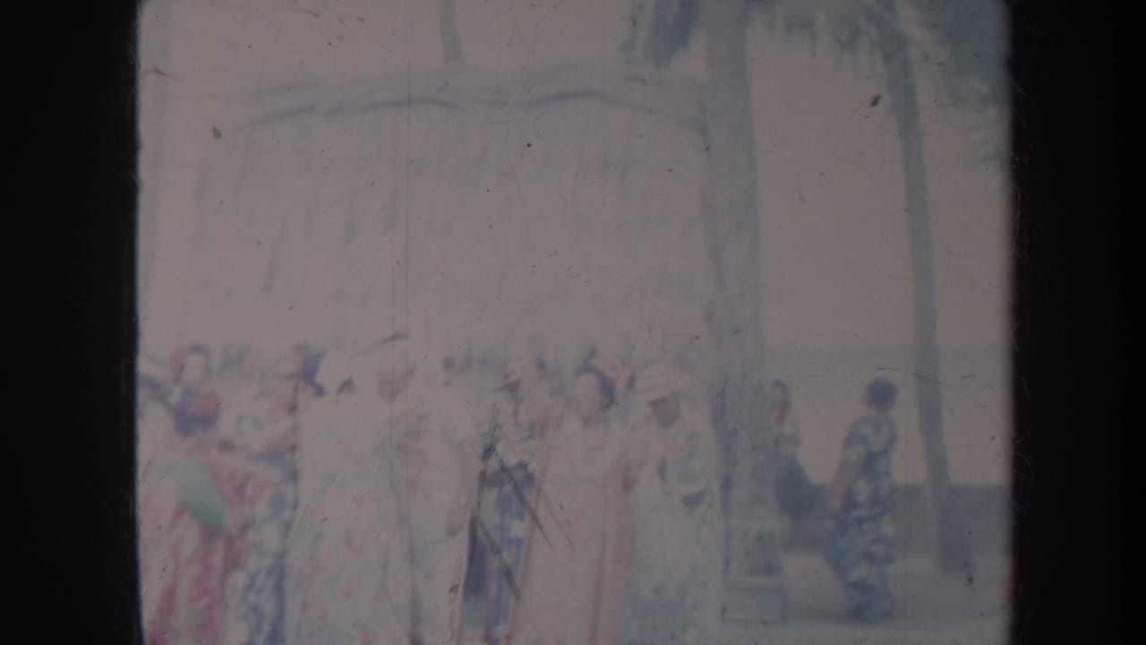 1967 Honolulu home-movie frame showing women in colorful floral gowns and hats gathered outdoors near palm trees.