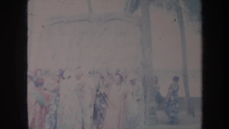 1967 Honolulu home-movie frame showing women in colorful floral gowns and hats gathered outdoors near palm trees.