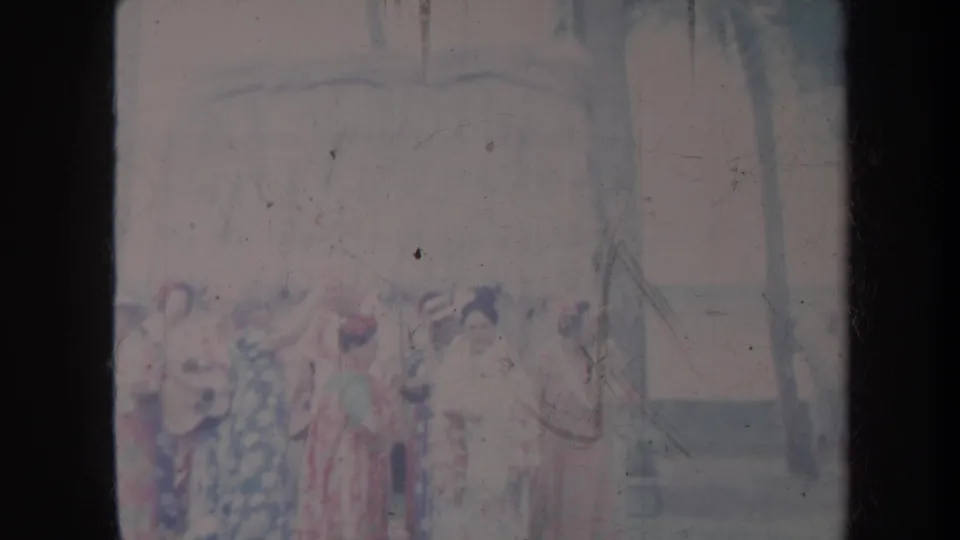 1967 Honolulu home-movie frame showing women in floral dresses gathered outdoors beside palm trees and a low structure.