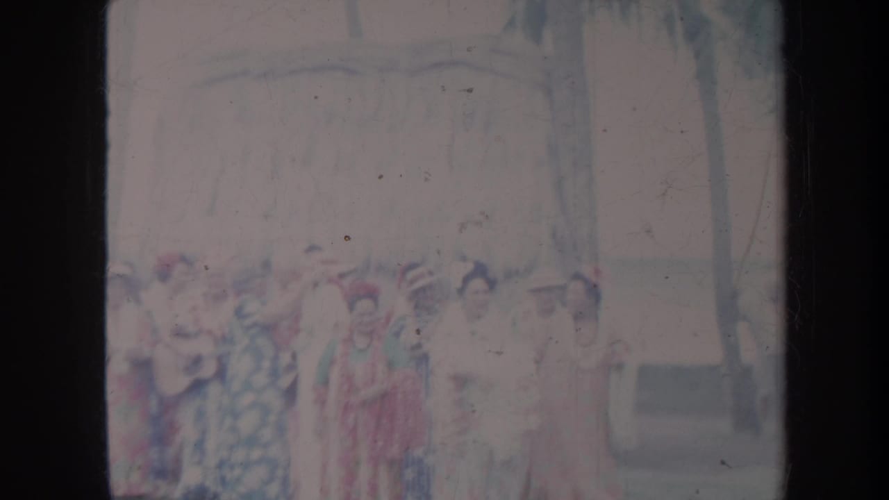 1967 Honolulu home-movie frame showing women in patterned dresses and hats gathered outdoors near a building and trees.