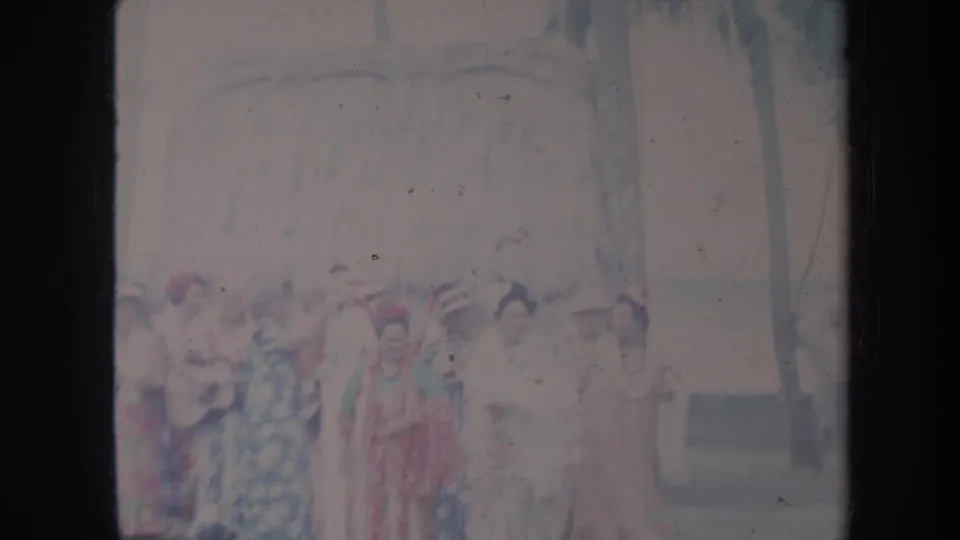 1967 Honolulu home-movie frame showing women in patterned dresses and hats gathered outdoors near a building and trees.