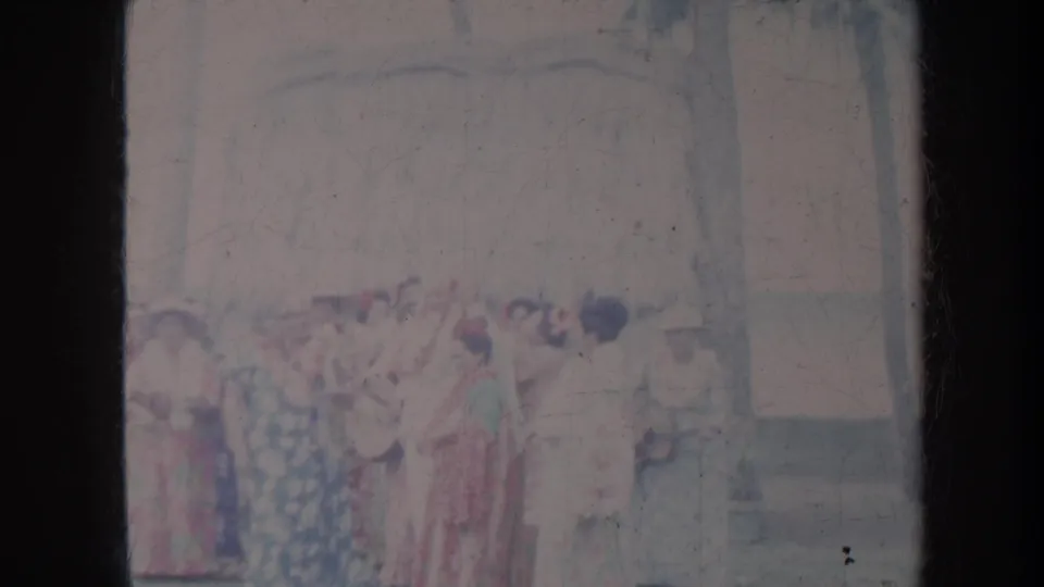 1967 Honolulu 8mm frame showing women in patterned dresses grouped outdoors, with a figure at right near a building wall.