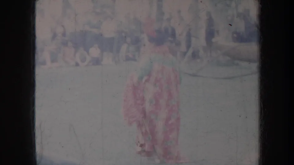 Faded Honolulu 8mm frame with a woman in a red patterned dress dancing outdoors as spectators watch in the background.