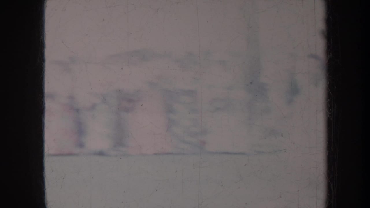 Soft, distant view of a crowd near a pale waterfront area, partly obscured by dark film borders.