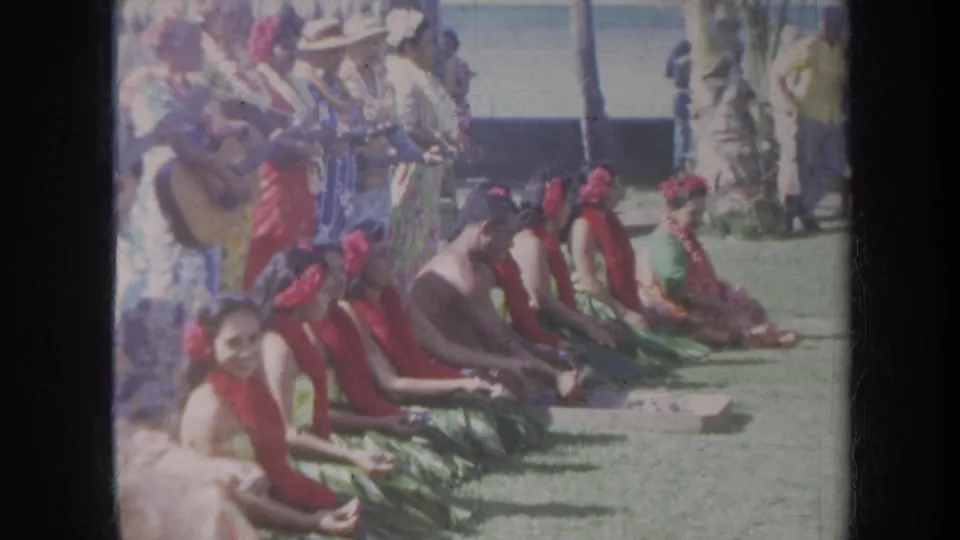 1967 Honolulu home-movie frame showing seated hula performers in red leis while musicians play behind them near palm trees and ocean.