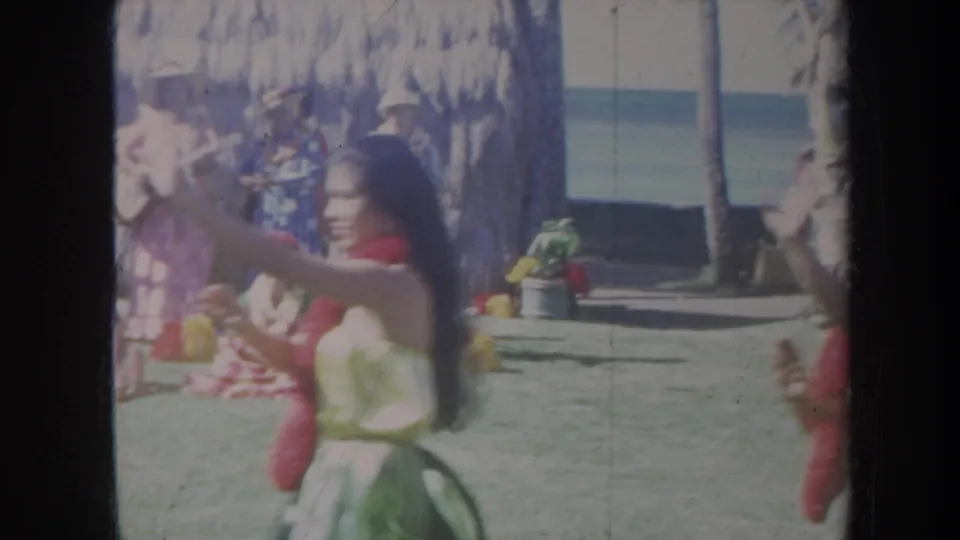 1967 Honolulu home-movie frame showing a hula dancer in a red lei moving before seated musicians and performers near the shore.