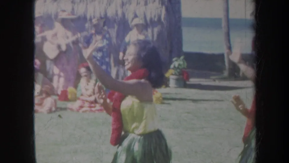 1967 Honolulu frame showing a smiling hula dancer gesturing in front of musicians, seated performers, palm trees, and ocean.
