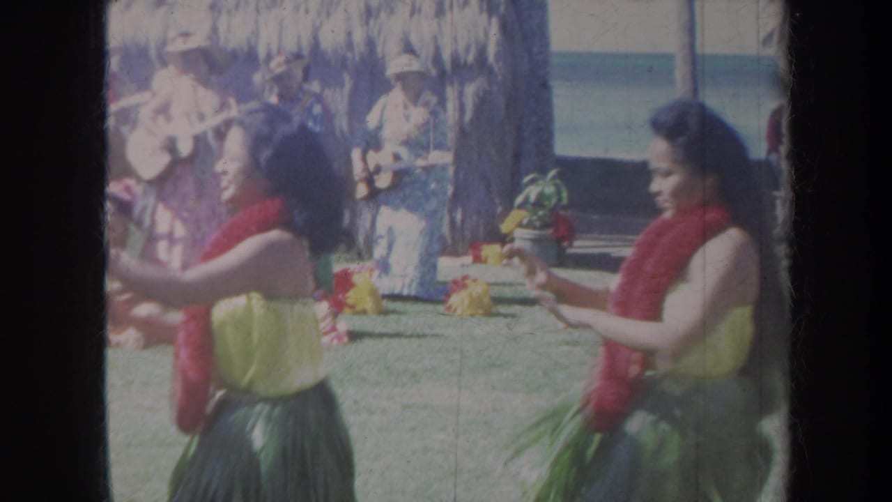 Hula dancers in red leis perform on a lawn with musicians and a thatched shelter behind them.