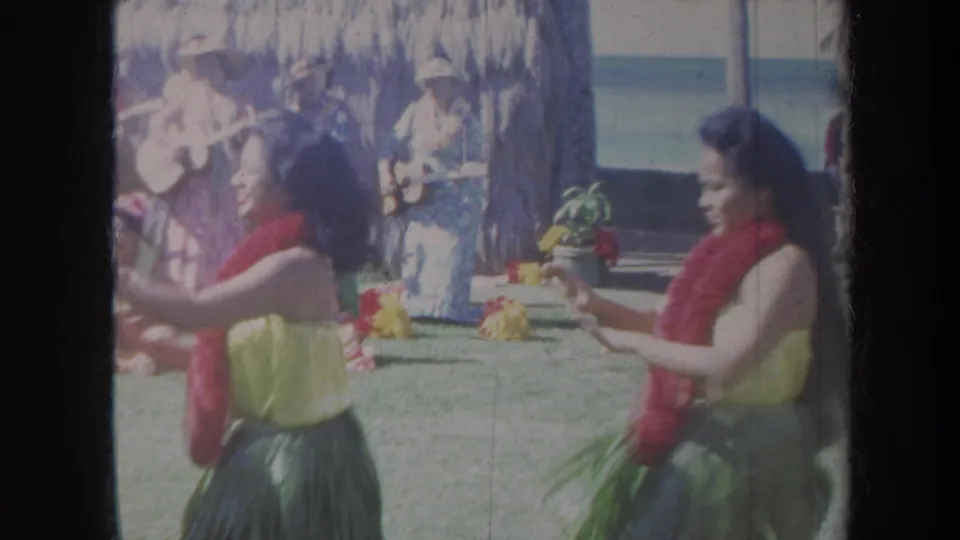 1967 Honolulu home-movie frame showing hula dancers in leis and grass skirts performing outdoors near musicians and palm trees.