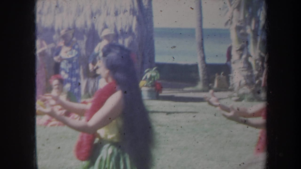 A hula dancer in a red lei moves across a lawn with the ocean and palm trees in the background.